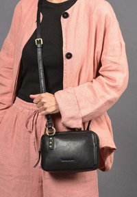 Bolso cruzado de cuero negro con forma rectangular, cierre de cremallera y detalles decorativos en el hardware. Llevado con una chaqueta de lino rosa.