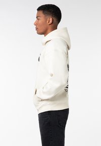 Cream hoodie with a front kangaroo pocket, black graphic prints on chest and sleeve, ribbed cuffs, and hem; paired with black jeans.