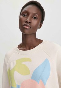 Vaalean beige-sweater, jossa on pyöreä kaula-aukko, ja siinä on abstrakteja kukkakuoseja vihreänä, sinisenä ja vaaleanpunaisena. Materiaali on pehmeää puuvillaa.