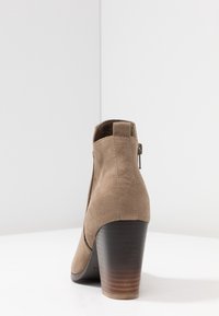 Beżowy zamszowy botek z zaokrąglonym noskiem i obcasem na słupku. Posiada eksponowany zamek błyskawiczny oraz gładką, teksturowaną powierzchnię.