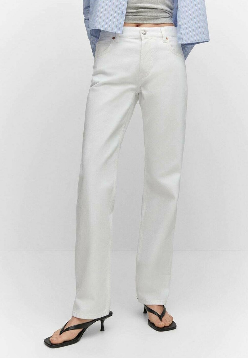 Personne portant un pantalon blanc droit taille haute associé à des sandales noires à petit talon et un top court gris sous une chemise bleue rayée.