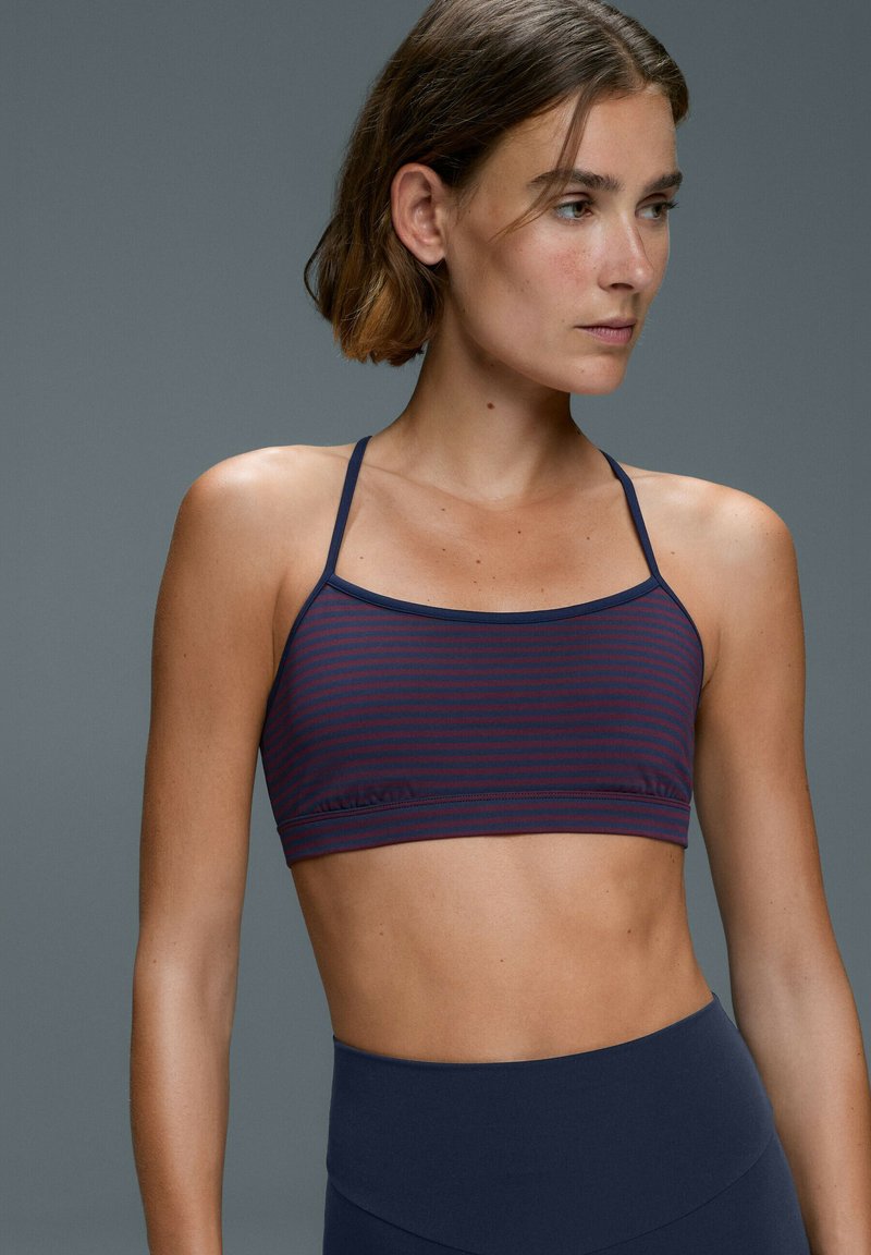Femme aux cheveux courts portant un soutien-gorge de sport rayé bleu marine et bordeaux et un legging taille haute bleu marine sur un fond gris uni.
