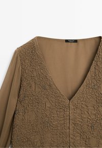 Blouse brun clair texturée avec un décolleté en V profond et de petites décorations perlées à l'avant et des manches longues.