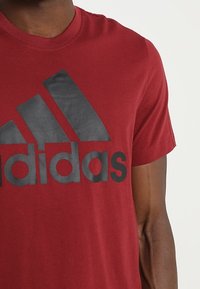 Punainen puuvillainen T-paita, jossa on musta Adidas-logo ja kolme vinoviivaa. Lyhyet hihat, pyöreä kaula-aukko. Pehmeä tekstuuri.