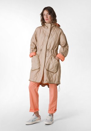 Beige gewatteerde parka met elastische taille en oranje manchetten, gecombineerd met koraalkleurige broeken en witte sneakers met blauwe accenten. Smeuïge textuur.