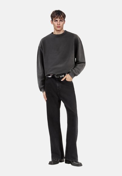Grijze oversized sweatshirt met ronde hals, gecombineerd met zwarte brede corduroy broeken. Model staat tegen een effen achtergrond.