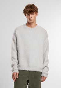 Hellgrauer, fusseliger Pullover mit rundem Hals und geripptem Saum, mit langen Ärmeln und lockerer Passform. Über olivgrünen Hosen getragen.