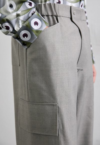 Graue maßgeschneiderte Hose mit strukturierter Oberfläche, ausgestattet mit Seitentaschen und einem flachen Design. Kombiniert mit einem gemusterten Hemd.