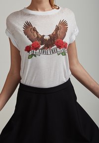 Hvid T-shirt med et ørne grafik, røde roser og en bannertekst "STONE FREE." Casual pasform med korte ærmer. Sort nederdel nedenunder.