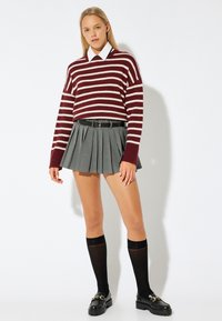 Maglione a righe bordeaux e crema, gonna plissettata grigia, cintura nera, calze nere fino al ginocchio, mocassini neri chunky. Tessuti dalla trama liscia.