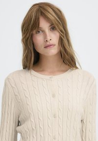 Beige kabelstrik cardigan med rund halsudskæring og lukning med fem knapper. Har et struktureret mønster og bløde, ribbede manchetter.