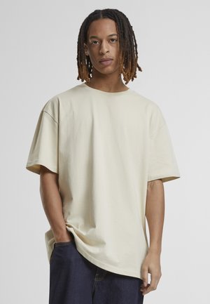 Ung person med mellemlange dreadlocks, iført en løs off-white T-shirt og mørkeblå jeans, stående med den ene hånd i lommen.