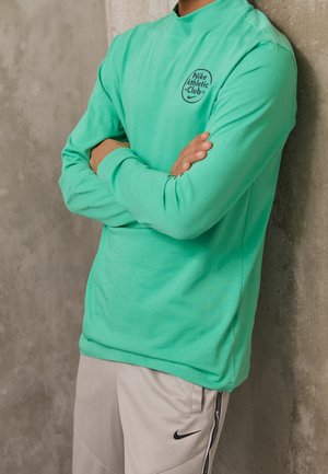Personne portant une chemise à manches longues verte avec le logo "Nike Athletic Club" et un pantalon beige avec le swoosh noir de Nike, les bras croisés contre un mur en béton.