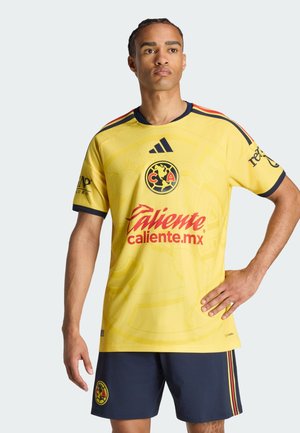 Mann trägt gelbes Club América Fußballtrikot und marineblaue Shorts mit Logos, steht mit einer Hand auf der Hüfte vor schlichtem hellen Hintergrund.