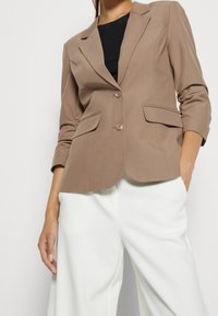 Brun skräddarsydd kavaj med en enkel knappstängning, två framfickor och trekvartslånga ärmar, i kombination med vita vida byxor.