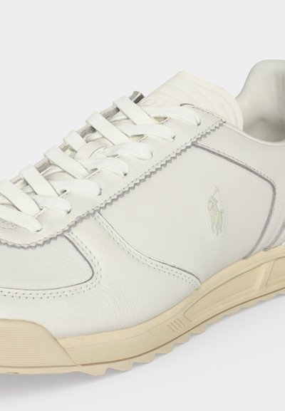 Άσπρο δερμάτινο αθλητικό παπούτσι με λευκά κορδόνια, κρεμ σόλα και ανάγλυφο λογότυπο στο πλάι, εμφανιζόμενο σε ελαφρώς γωνιακή θέση.