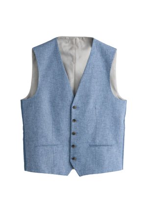 Mladý muž v modrom trojdielnom obleku, bielej košeli, vzorovanej čiernej kravate, čiernych topánkach, stojaci s rukami v vreckách oproti béžovej stene.