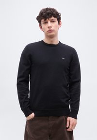 Jeune homme aux cheveux courts et bouclés portant un pull noir Levi's et un pantalon marron, debout avec une main dans la poche devant un fond uni.