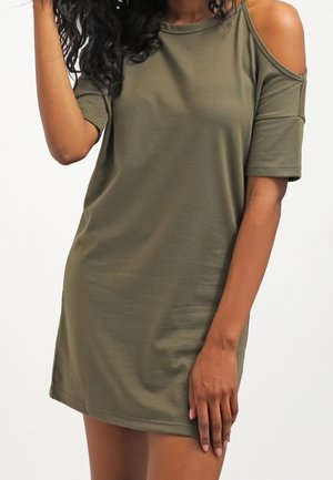Kobieta w krótkiej oliwkowozielonej sukience z wycięciem na zimne ramię na prawej ręce, na jednolitym białym tle.