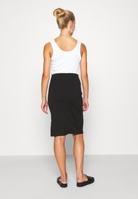 Jupe midi noire avec une coupe ajustée, associée à un débardeur blanc. Le mannequin a une coiffure lisse et porte des chaussures noires à enfiler.