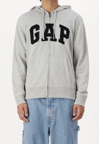Grauer Hoodie mit Reißverschluss und schwarzem "GAP"-Logo in fetten Buchstaben, aus weichem Stoff, mit Kapuze und Fronttaschen.