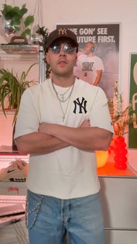 Bílá tričko s černým logem New York Yankees, modré džíny, stříbrné řetízkové akcenty, sluneční brýle a tmavá baseballová čepice. Vnitřní prostředí, viditelné rostliny.