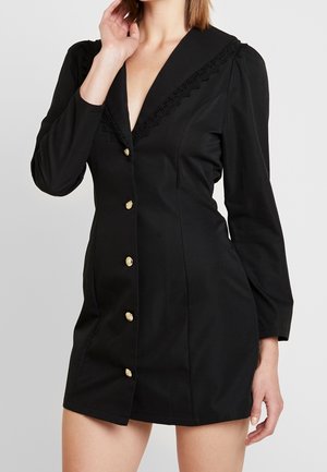 Femme portant une mini robe noire ajustée à manches longues, avec des boutons dorés à l'avant et un décolleté en V orné de dentelle, main près du visage.