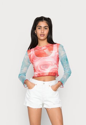 Top cropped de manga longa com estampa floral em rosa e azul. O tecido é transparente com bordas escallopadas vermelhas. Combinado com calções brancos.