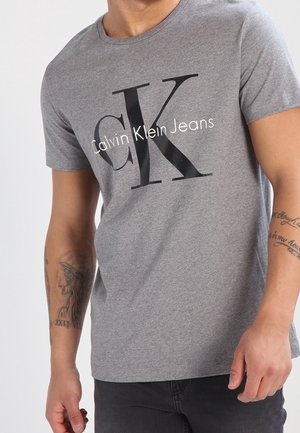 Mężczyzna noszący szarą koszulkę Calvin Klein Jeans z czarnym logo i napisem, pokazujący tatuaże na obu przedramionach.