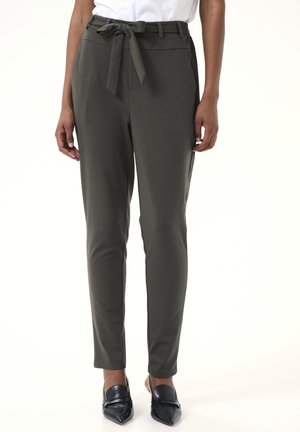 Dark green tailored trousers with a smooth texture, a tied belt detail at the waist, and side pockets. Paired with pointed metallic shoes.