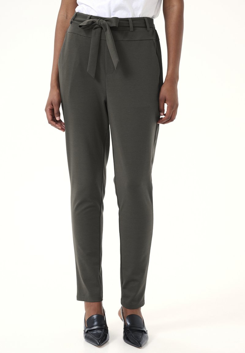 Pantaloni su misura verde scuro con una texture liscia, dettaglio di cintura legata in vita e tasche laterali. Abbinati a scarpe metalliche a punta.