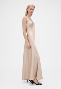 Robe maxi en satin beige avec fines bretelles, encolure incurvée et texture lisse. Associée à des talons ouverts noirs.