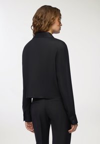 Giacca nera cropped con maniche lunghe, tessuto liscio, colletto classico e polsini con bottone. Vestibilità tailored, design minimalista.