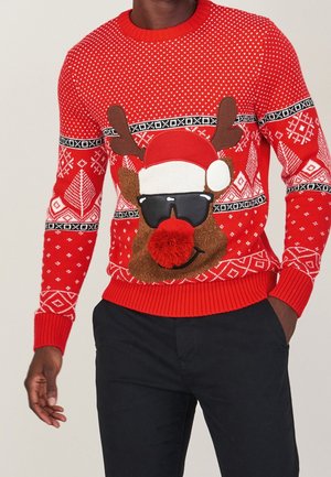 Pull de Noël rouge avec des motifs blancs et un renne portant des lunettes de soleil, un chapeau de Père Noël et un gros pompon rouge sur le nez.