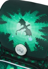 Zielony plecak z sylwetką smoka na tle lasu, z efektami świetlnymi w kształcie gwiazd i okręgów; zawiera okrągły detal logo.