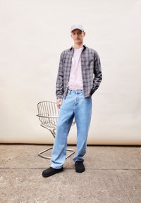 Grijze geruite shirt over een lichtroze overhemd met knopen, gecombineerd met lichtblauwe losse spijkerbroek en zwarte instap schoenen. Eenvoudige grijze pet. Metalen stoel op de achtergrond.