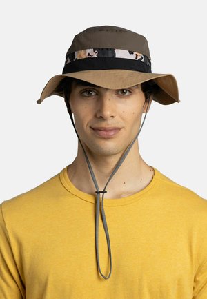 Hombre con sombrero tipo bucket marrón y negro con correa para la barbilla y camiseta amarilla con cuello redondo, mirando hacia adelante con expresión neutral.