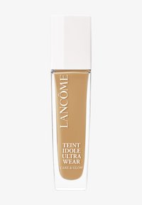 Grunnleggende flaske med en transparent firkantet bunn og et hvitt firkantet lokk. Brun væske inni, merket "Teint Idole Ultra Wear Care & Glow."
