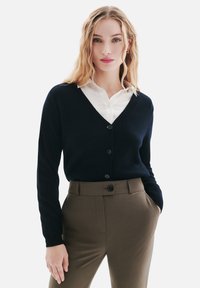 Cardigan bleu marine avec un col en V, des manches longues et des boutons à l'avant, associé à une chemise blanche à col et un pantalon olive ajusté.