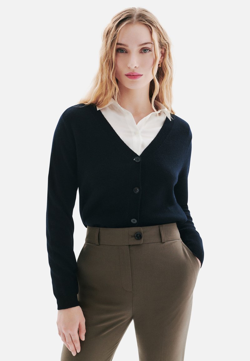 Cardigan bleu marine avec un col en V, des manches longues et des boutons à l'avant, associé à une chemise blanche à col et un pantalon olive ajusté.