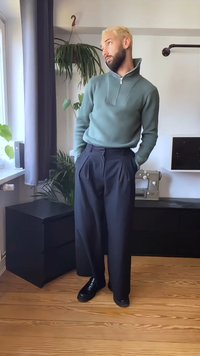 Green zip-up sweater with a high collar, paired with dark pleated trousers. Model stands with hands in pockets on wooden floor near plants.