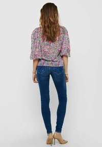 Bluse mit Blumenmuster und Puffärmeln, kombiniert mit blauen Skinny-Jeans und nude-farbenen High Heels. Rückansicht vor einem neutralen Hintergrund präsentiert.