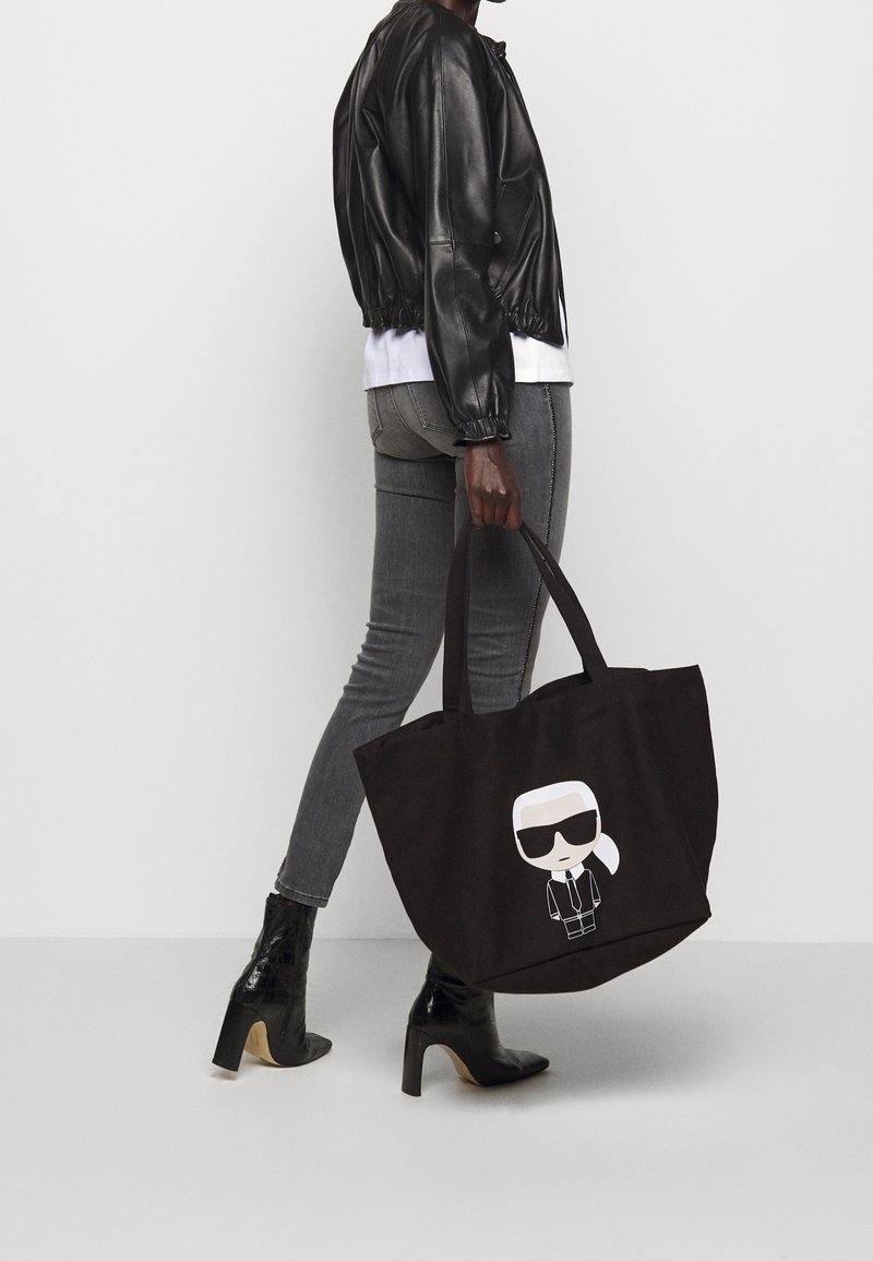 Personne portant une veste en cuir noire, un jean skinny gris et des bottes noires à talons, tenant un sac cabas noir avec un motif de personnage de dessin animé.