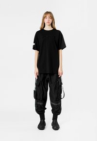 Camiseta negra de gran tamaño con el logotipo "MWM" de cuentas, combinada con pantalones cargo negros holgados que tienen múltiples bolsillos y detalles en metal.