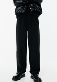 Personne portant un pantalon noir large, des chaussures en cuir noir et une veste en cuir noir avec une fermeture éclair, se tenant devant un fond blanc.