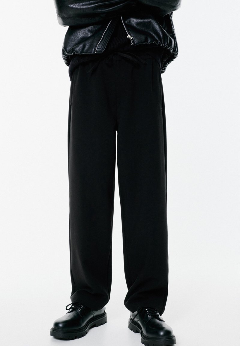 Personne portant un pantalon noir large, des chaussures en cuir noir et une veste en cuir noir avec une fermeture éclair, se tenant devant un fond blanc.