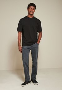 Man staat en lacht, draagt een zwart T-shirt, grijze spijkerbroek en zwarte sneakers met witte zolen op een effen lichte achtergrond.