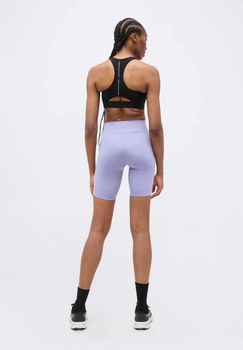 Vrouwelijke atleet staat met de rug naar de camera, draagt een zwarte sportbeha, lichtpaarse fietsbroek, zwarte sokken en grijze sneakers, tegen een effen achtergrond.