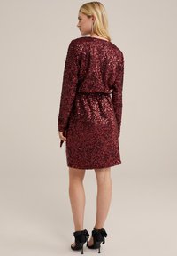 Rode paillettenjurk met lange mouwen en een broekriem in de taille. Glad textuur, kniehoogte, aangevuld met zwarte hoge hakken met strikken.