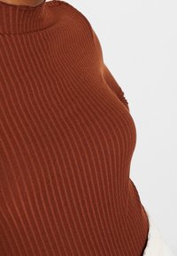 Gros plan d'une personne portant un pull col roulé marron ajusté à côtes, sur un fond clair uni.